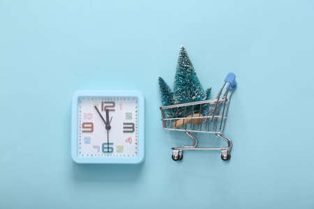 christmas composition. Alarm clock and shopping trolley with mini Christmas trees on blue background