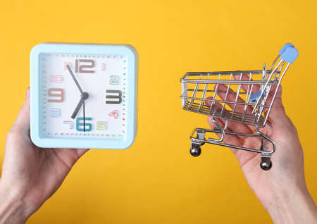 Hands holding alarm clock and shopping trolley on yellow background. shopping timeの写真素材