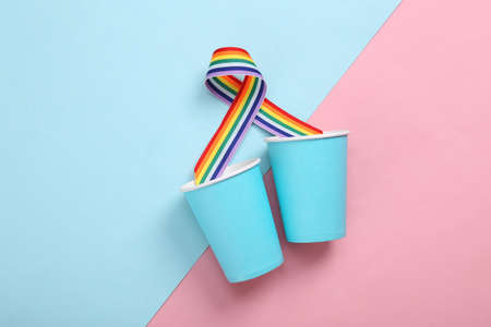 Paper cups with LGBT rainbow ribbon pride tape symbol on pink blue background.