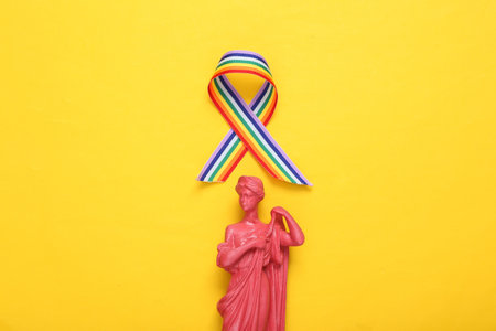 Ancient Greek goddess statue with LGBT rainbow ribbon pride tape symbol on yellow background. top viewの写真素材