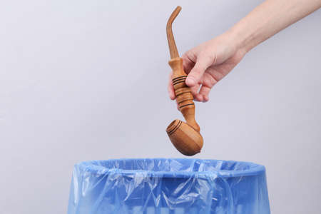 Hand throws smoking pipe into trash bin with package on gray background
