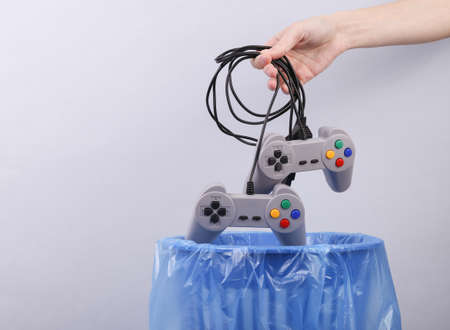 Hand throws plastic gamepads into trash bin with package on gray background