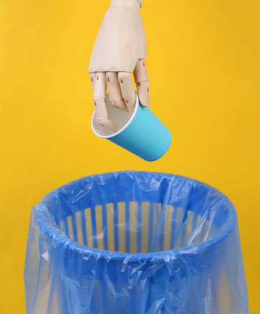 Wooden hand throws paper cup into trash bin with package on yellow background