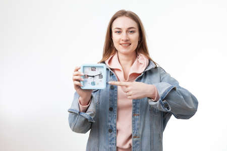 Young smiling woman holding clock and points finger at it isolated on white backgroundの写真素材