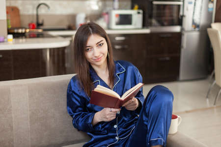 Young woman in nightie reading a book while sitting on the sofa in the living room