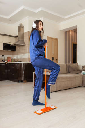 Cheerful young woman in nightie washes the floor with a mop and listens to music on headphones in...