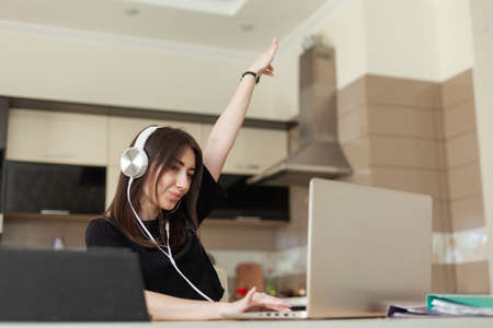Young woman looking into a laptop and listening to music on headphones while sitting at a table in a...