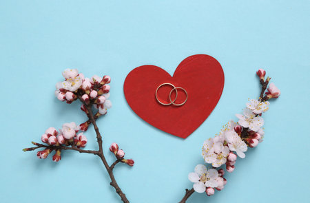 Wedding, spring concept. Red heart with wedding rings and flowering branches on a blue backgroundの写真素材