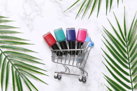 Mini shopping cart with nail polish bottles on marble background with palm leaves