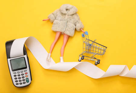 Woman Doll in fur coat with shopping trolley on payment tape of pos terminal. Minimalist shopping...