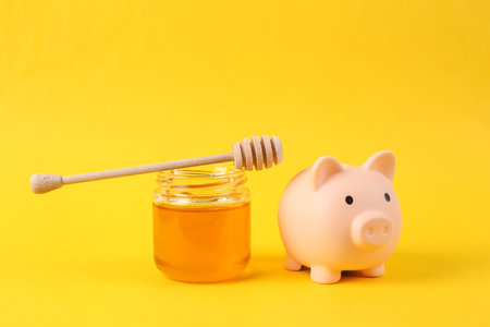 Piggy bank and Bee honey jar with honey wooden spoon on yellow backgroundの写真素材