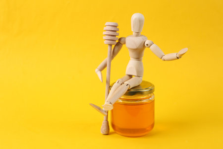 Wooden puppet holds honey spoon and sits on honey jar on yellow backgroundの写真素材