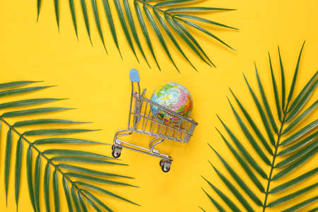 Shopping trolley with globe and palm leaves on yellow background. Tropical, minimal layout. top view. flat layの写真素材