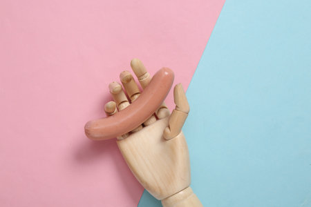 Wooden hand holds a sausage on pink blue background. Minmalistic food concept. flat lay
