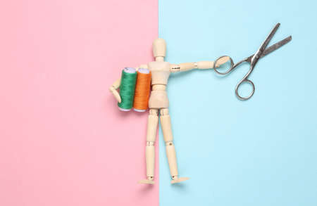 Wooden puppet holding Skeins of colored thread and scissors on a blue-pink pastel background. hobby