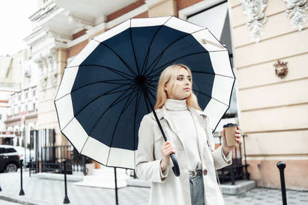 Young blonde woman in trench coat with umbrella and coffee cup in the city