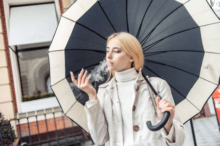Young blonde woman smokes electronic cigarette under umbrella in the cityの写真素材