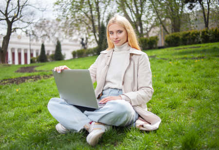 Young blonde woman uses a laptop while sitting on the grass in the park