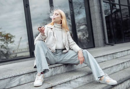 Young stylish blonde woman smokes electronic cigarette while sitting on stairs near business center