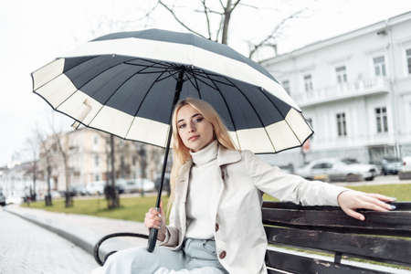 Young blonde woman sitting on a bench with umbrella in cityの写真素材