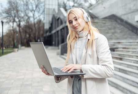 Young blonde woman in headphones holding laptop outdoorsの写真素材