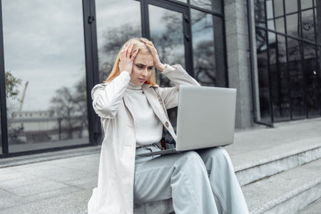 Young woman holding a headache and sitting with a laptop on the stairs near the business center Headache, negative emotionsの写真素材