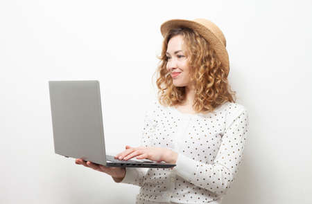 Cute smiling curly-haired caucasian woman in straw hat uses laptop on white backgroundの写真素材