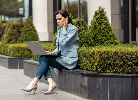 Cute Caucasian woman is using laptop in the city outdoors. Distant work. Modern business woman....