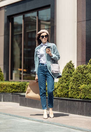 Stylish shopaholic woman in trendy trench coat, beret, sunglasses, high heels and jeans walks...