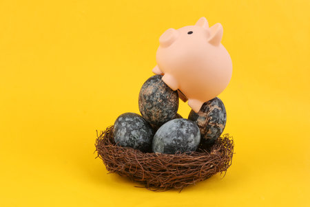 Piggy bank sitting on Gray marble painted Easter eggs in nest on yellow backgroundの写真素材