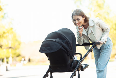 Young smiling mom calming baby in stroller showing hush gesture in parkの写真素材