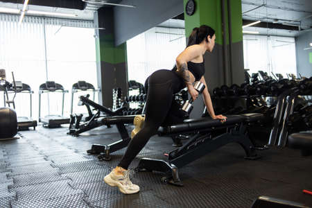 Fitness woman doing dumbbell rows with one hand leaning on a bench in the gymの写真素材