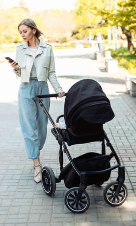 Stylish mom with stroller using smartphone in autumn parkの写真素材