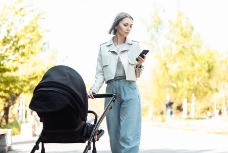 Stylish mom with stroller using smartphone in autumn parkの写真素材