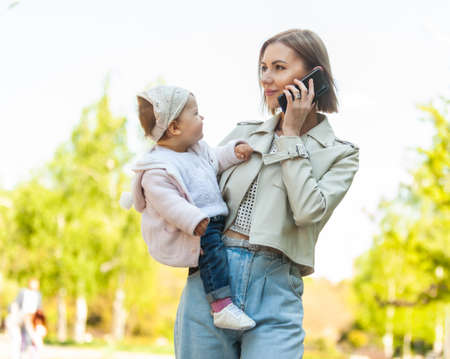 Stylish young mother with daughter in her arms talking on the phone in the parkの写真素材