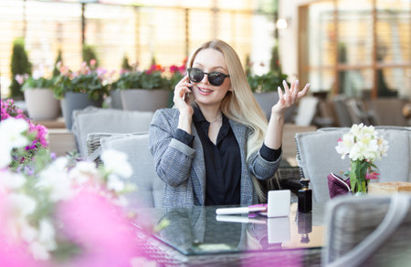 Young blonde woman in sunglasses talking on the phone while sitting at a table in the summer terrace of cafeの写真素材