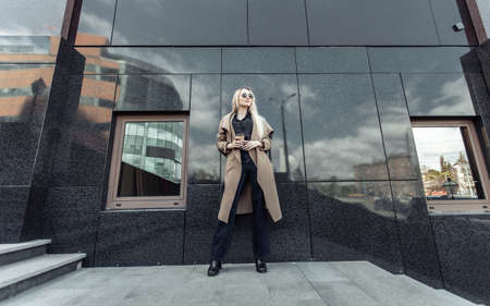 Blonde woman in trench coat holds cup of coffee in urban locationの写真素材