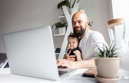 Happy bearded working father with little daughter uses laptop while sitting at table in modern roomの写真素材