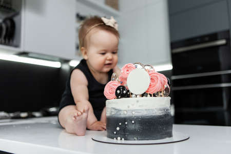 Lovely little baby girl is sitting at the table with a birthday cake in the kitchen.の写真素材