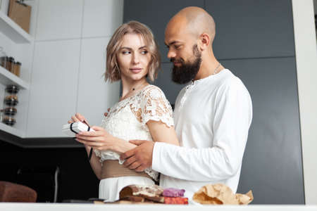 Handsome man makes a surprise to his lovely woman while she is cooking in the kitchen. The husband gives a box with a gift to his wife. romantic love concept. birthday or anniversaryの写真素材
