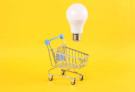 Shopping trolley with floating light bulb on yellow bright background. Minimal shopping concept