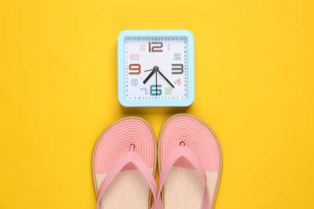 Colored flip flops with alarm clock on yellow background. Beach vacation concept. flat lay