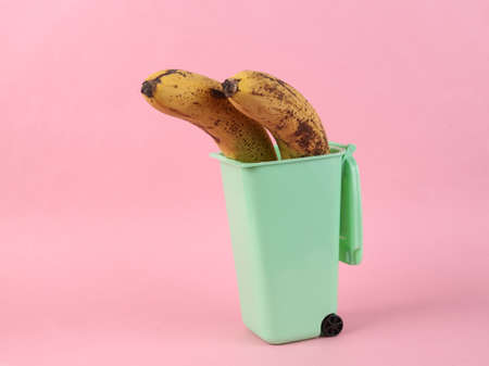 Spoiled bananas in mini trash can on pink background