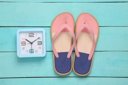 Colored flip flops with alarm clock on blue wooden background. Beach vacation concept. Travel time. flat layの写真素材