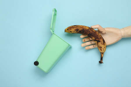 Recycling concept. Wooden hand throws rotten banana into the trash can on pink backgroundの写真素材
