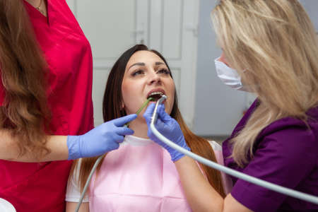 Female dentist with an assistant uses a dental drill in a dental clinic. Young woman patient at the...