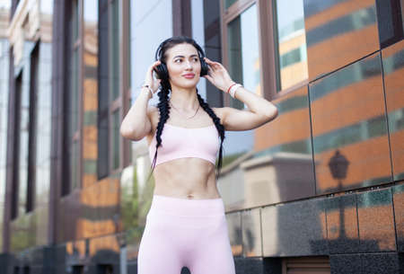 Portrait of a cute fit brunette woman with pigtails and headphones on the background of a city...