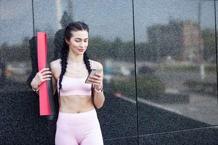Fit woman looking at the smartphone screen leaning against the wall of the building