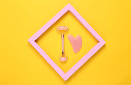 Face roller and gua sha stone massager in pink frame on yellow background. top view