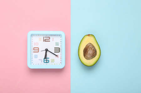 Avocado fruit and alarm clock on blue pink pastel background. top view